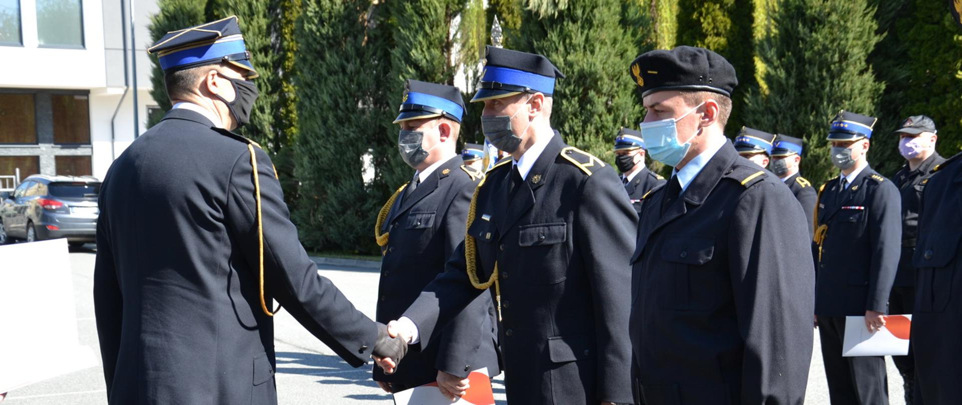Przed frontem szyku zbórki Komendant Powiatowy składa gratulacje trzem awansowanym podoficerom. Wszyscy w mundurach galowych . W tle zbiórka strażaków.