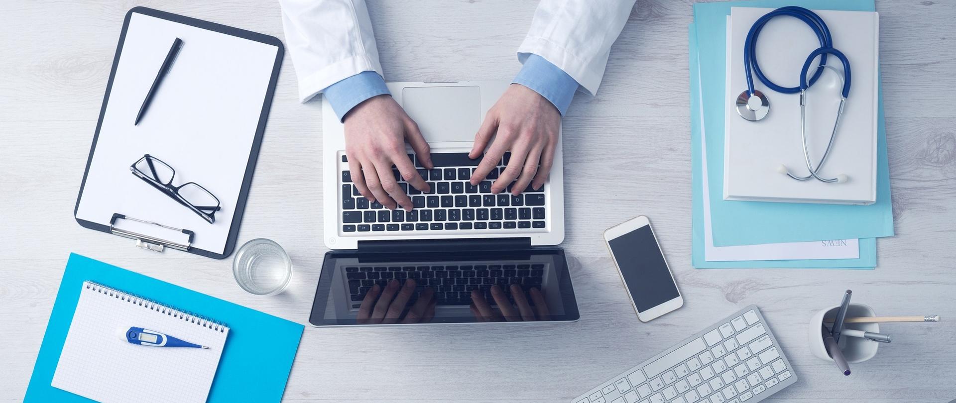 Komputer na biurku obsługiwany przez lekarza. Obok notatnik, termometr, okulary, klawiatura, stetoskop i smartfon.