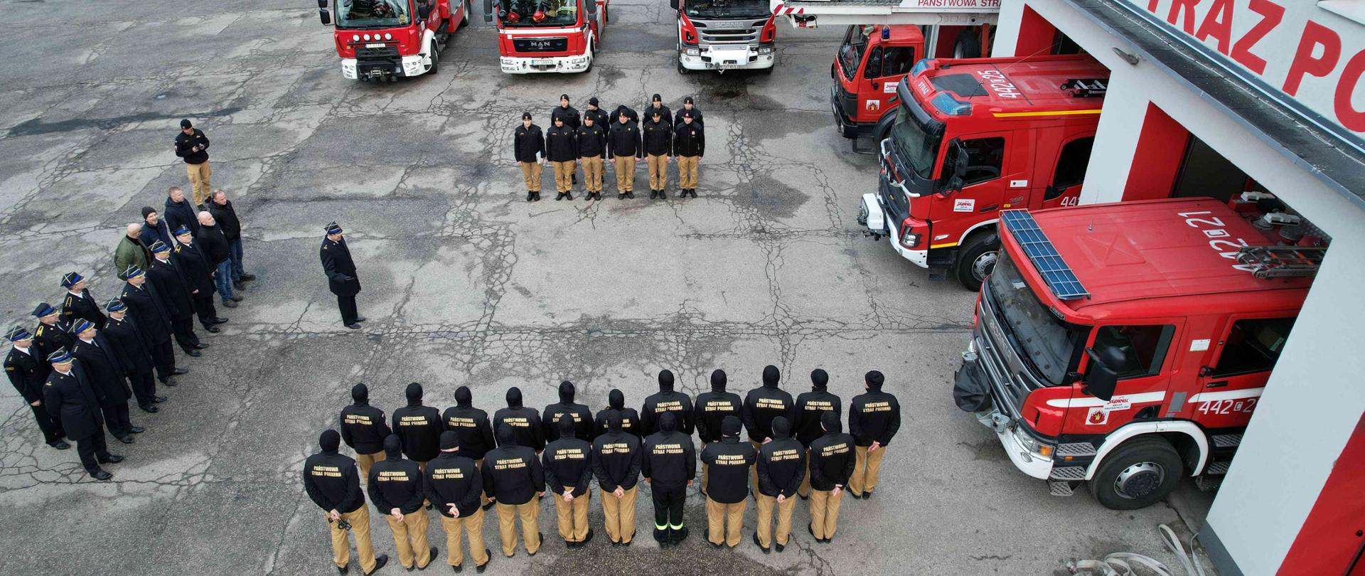 Zbiórka strażaków na tle czerwonych wozów bojowych widziana z lotu ptaka.