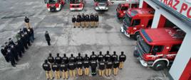 Zbiórka strażaków na tle czerwonych wozów bojowych widziana z lotu ptaka.