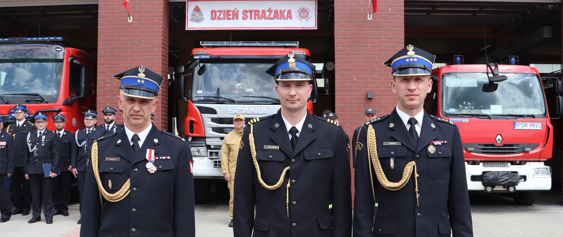 trzech strażaków stoi obok siebie, w tle garaże z samochodami strażac kimi i grupa strażaków