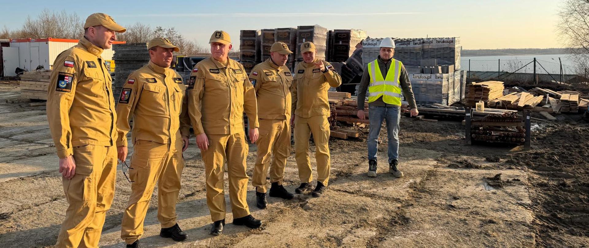 Przedstawiciele kierownictwa PSP podczas wizyty na terenie budowy Podkarpackiego Centrum Ratownictwa w Tarnobrzegu, stojący obok materiałów budowlanych i infrastruktury placu budowy.