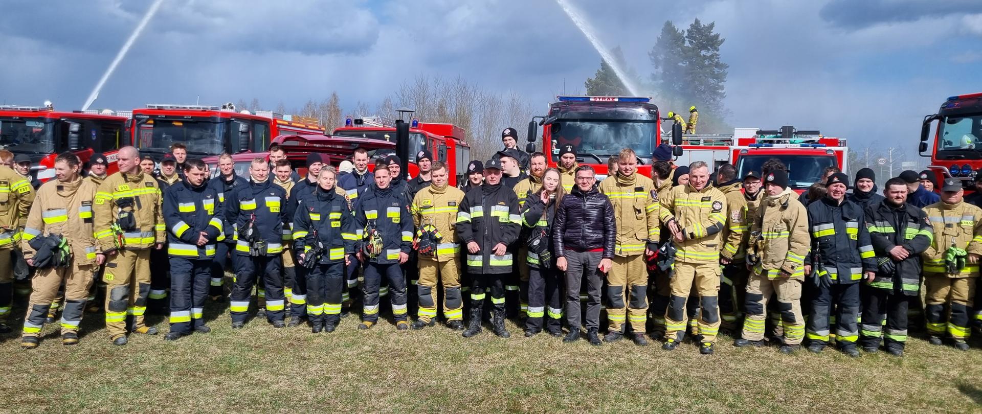Strażacy w jasnych i ciemnych mundurach na wspólnym zdjęciu po skończonych ćwiczeniach. Za nimi samochody pożarnicze i lejąca się w górę woda ze zwartego strumienia z działek wodno-pianowych samochodów. 