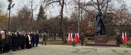 Żołnierze stoją obok pomnika Marszałka Józefa Piłsudskiego obok stoją flagi a w rzędzie przedstawiciele służb mundurowych oraz zaproszeni goście.