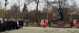 Żołnierze stoją obok pomnika Marszałka Józefa Piłsudskiego obok stoją flagi a w rzędzie przedstawiciele służb mundurowych oraz zaproszeni goście.