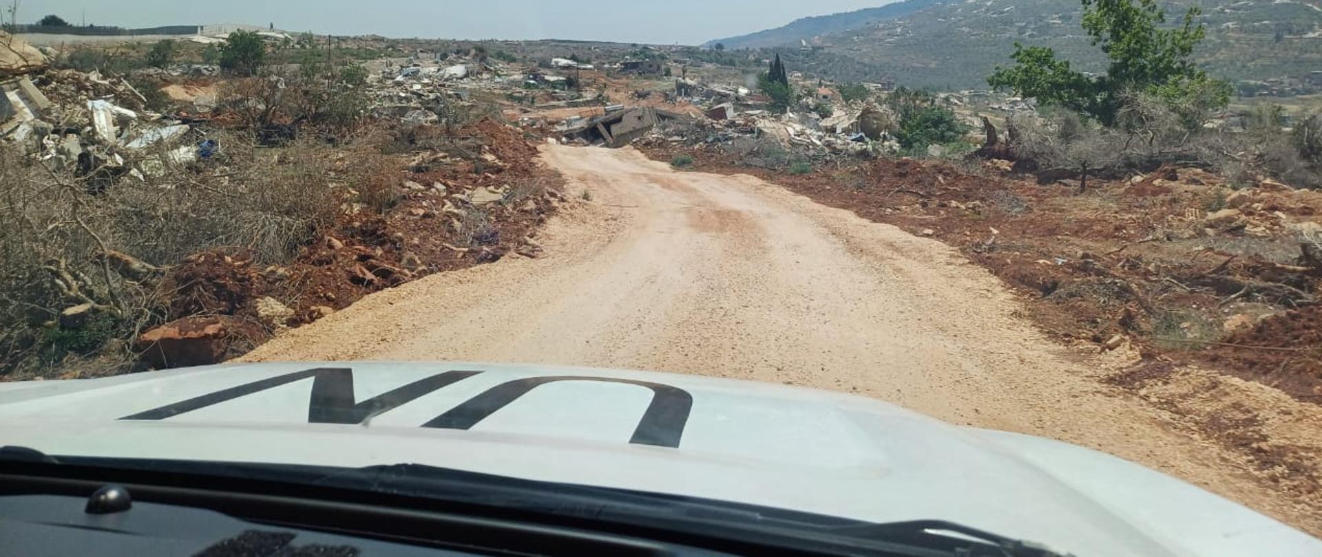 Samochód z oznaczeniem UN jedzie drogą, po boku zniszczone domy