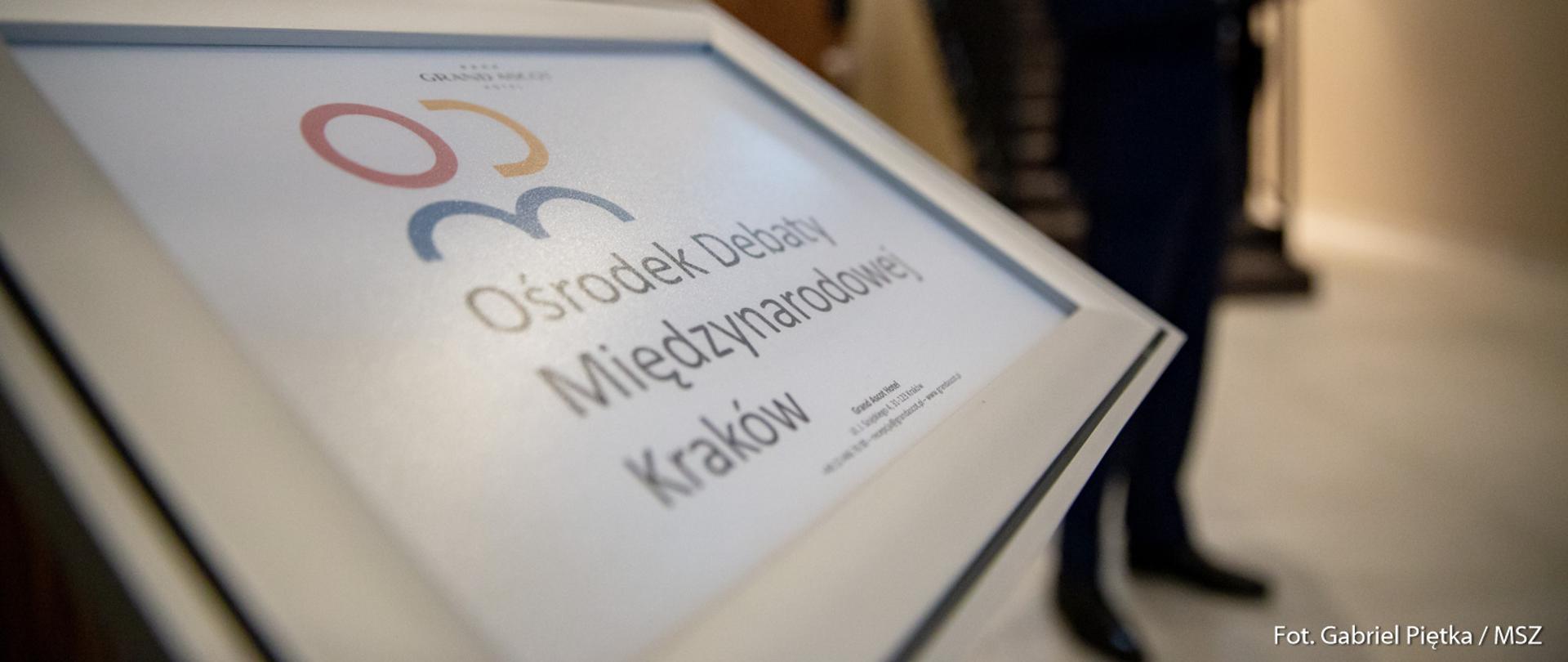 Ekran z logotypem Ośrodka Debaty Międzynarodowej w Krakowie, w tle niewyraźny człowiek w garniturze.