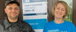 Na zdjęciu strażak stoi przy drzwiach wejściowych do punktu szczepień ubrany w czarny strój oraz pani pielęgniarka w niebieskiej koszulce oraz białych spodniach