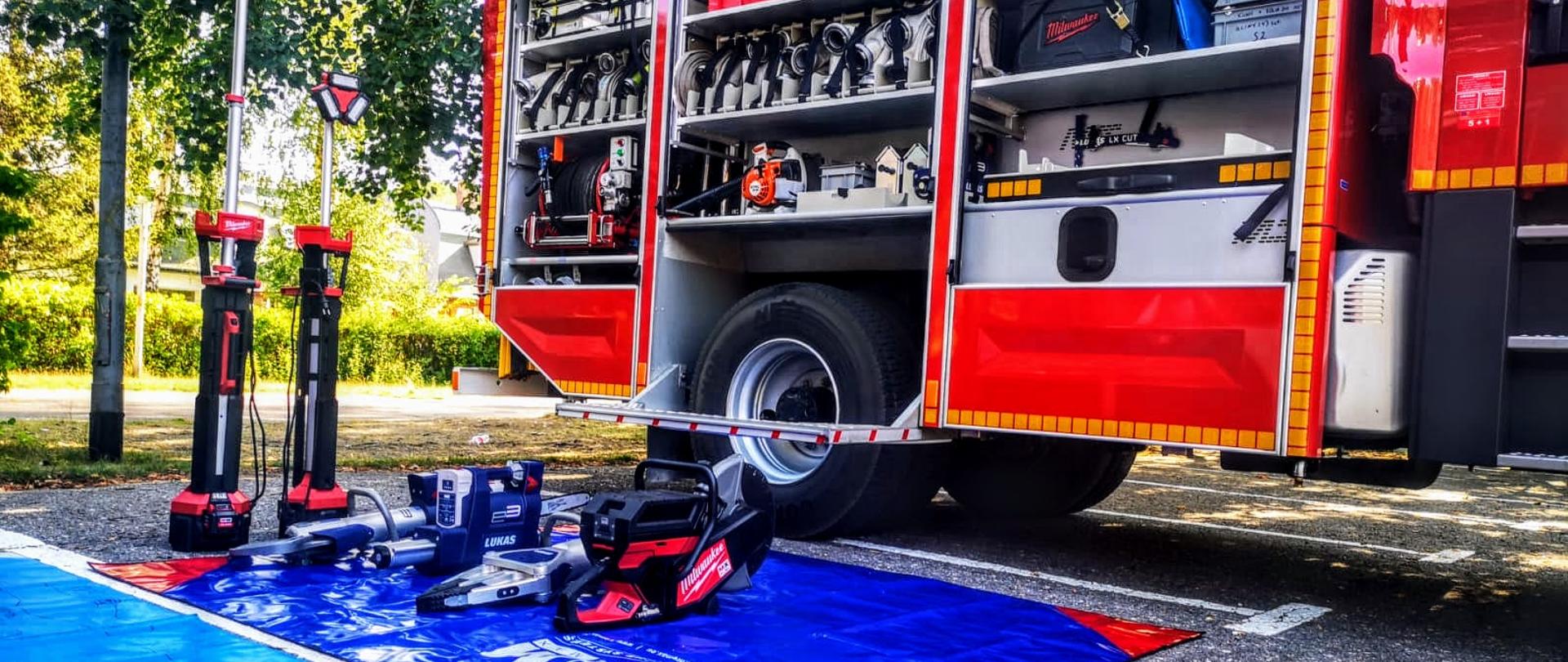 Zdjęcie przedstawia sprzęt hydrauliczny o zasilaniu akumulatorowym wraz z piłą tarczową oraz najaśnicami akumulatorowymi na niebieskiej plandece na tle czerwonego samochodu pożarniczego.