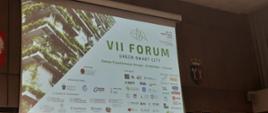 Widok na wielki ekran z zielonym napisem VII Forum Green Smart City na białym tle i ze zdjęciem budynku z balkonami obrośniętymi roślinami.