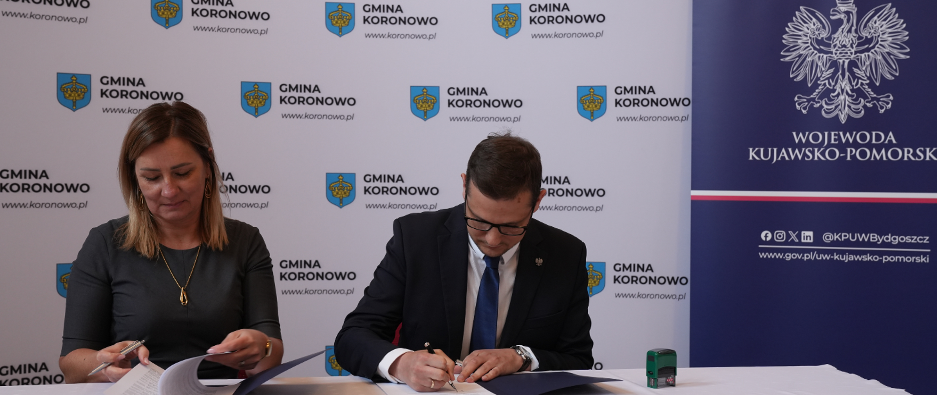 Podpisanie umowy przez wojewodę Michała Sztybla w powiecie bydgoskim. Wojewoda widoczny przy stole na tle ścianki gminy Koronowo.