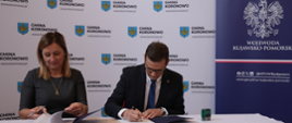 Podpisanie umowy przez wojewodę Michała Sztybla w powiecie bydgoskim. Wojewoda widoczny przy stole na tle ścianki gminy Koronowo.
