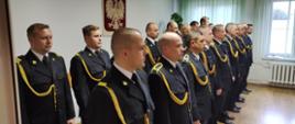 Zdjęcie przedstawia funkcjonariuszy podczas uroczystości mianowania na wyższe stanowiska służbowe. Strażacy stojący w dwuszeregu, znajdują się w świetlicy Komendy Powiatowej Państwowej Straży Pożarnej w Lwówku Śląskim. Przed strażakami znajduje się Komendant Powiatowy PSP w Lwówku Śląskim oraz Dowódca Jednostki Ratowniczo-Gaśniczej PSP w Lwówku Śląskim.