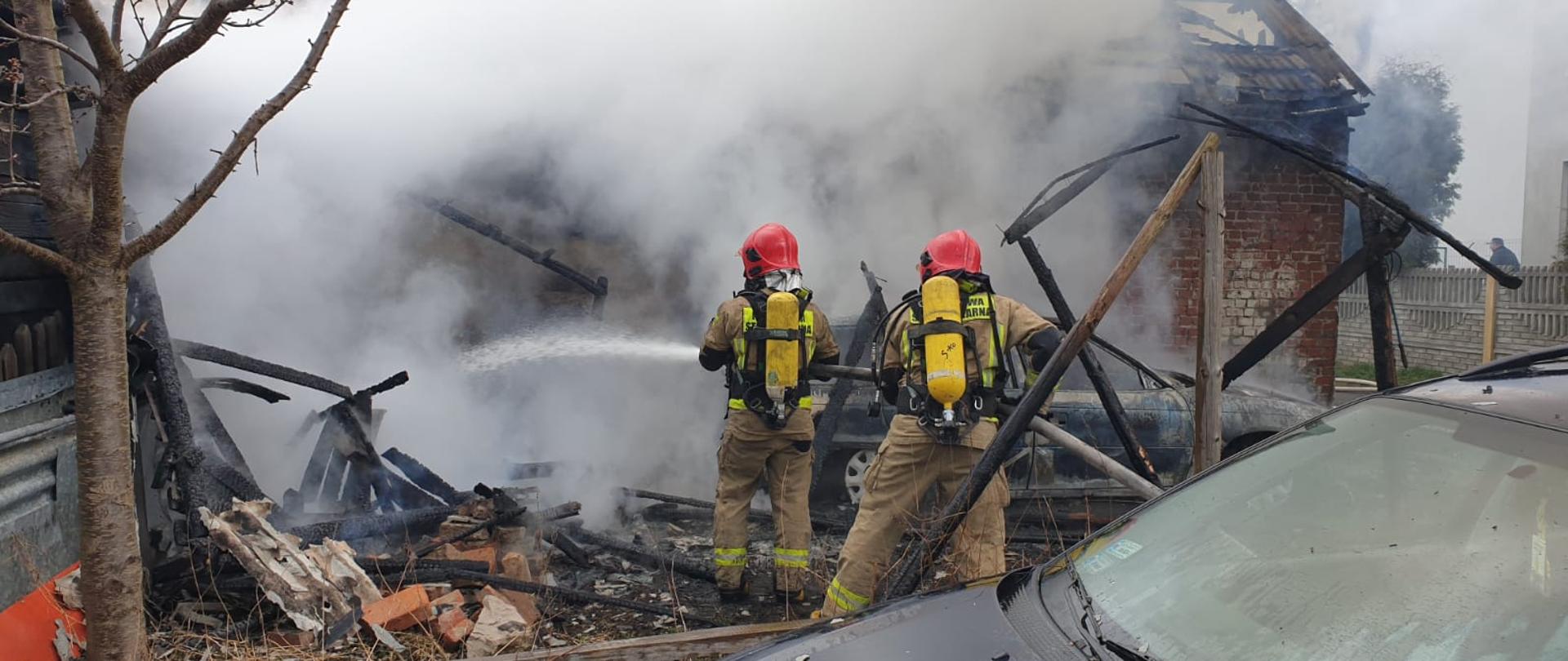 Na zdjęciu widać strażaków ubranych w ubrania bojowe i aparaty ochrony układu oddechowego, gaszących pożar budynku gospodarczego. Przed nimi widać gęste kłęby dymu. W pobliżu znajduje się samochód osobowy.