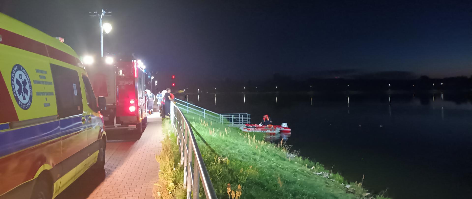 Zdjęcie przedstawia zalew w Strawczynie gdzie utonął młody mężczyzna, na wodzie unosi ponton Specjalistycznej Grupy Wodno-Nurkowej, którym działania prowadzili nurkowie. Po lewej stronie stoją samochody służb ratowniczych: ambulans i pojazd pożarniczy.