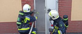 Zdjęcie przedstawia rotę strażaków w ubraniu specjalnym i SOUO oraz linią gaśniczą podczas wykonywania sekwencji wejścia do zadymionego pomieszczenia warsztatu JRG w Oleśnie. Z uchylonych drzwi wydobywa się sztuczny dym.