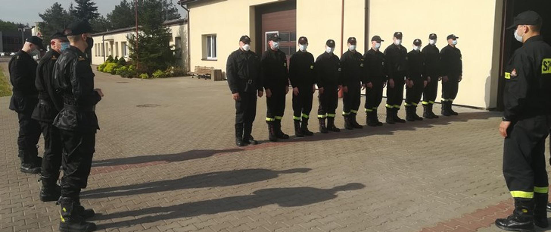 Zdjęcie przedstawia strażaków PSP stojących w szeregu przed budynkiem jednostki, z lewej strony stoją oficerowie PSP.
