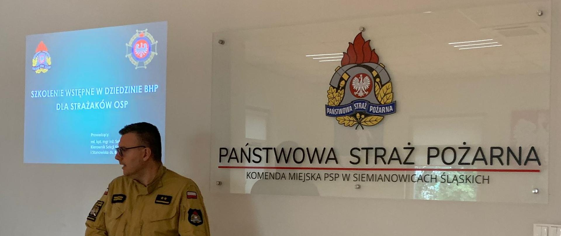 Mł. kpt. Sebastian Karpiński podczas zajęć prowadzonych podczas szkolenia podstawowego strażaków ratowników Ochotniczych Straży Pożarnych. Na tle od lewej strony treści szkolenia wstępnego BHP. Od prawej strony na ścianie logo PSP. 