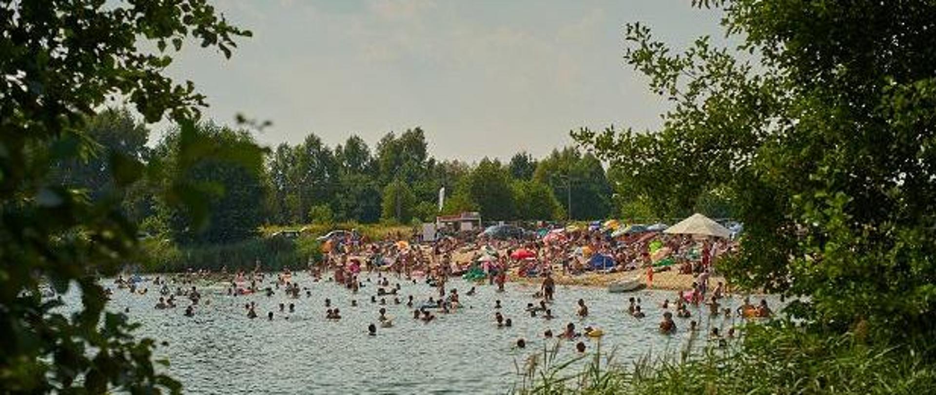 Ludzie kąpiący się nad jeziorem. Osoby w wodzie i na plaży. Widoczne parasole, materace, samochody. Wszystko otoczone roślinnością.