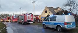Zastępy straży pożarnej stoją na drodze przed posesją. Z prawej strony stoi radiowóz policyjny. W głębi budynek mieszkalny