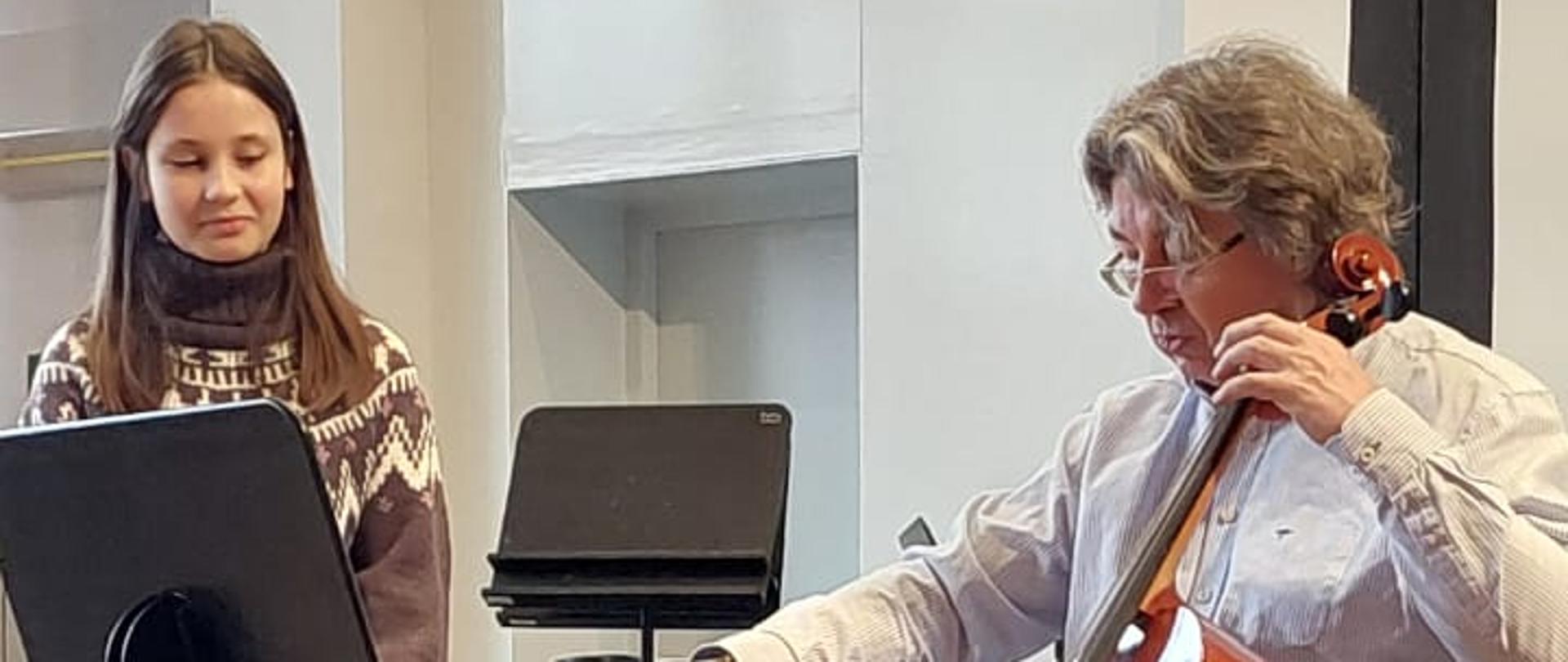 Mężczyzna gra na wiolonczeli , obok stoi dziewczynka i obserwuje.