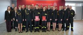 Grupa strażaków i pracowników Państwowej Straży Pożarnej stoi do wspólnego zdjęcia w sali konferencyjnej. W pierwszym rzędzie dwójka młodych strażaków w czarnych mundurach trzyma czerwone teczki z dokumentami. W tle znajduje się czerwony baner z napisem „Państwowa Straż Pożarna” i symbolem białego orła.