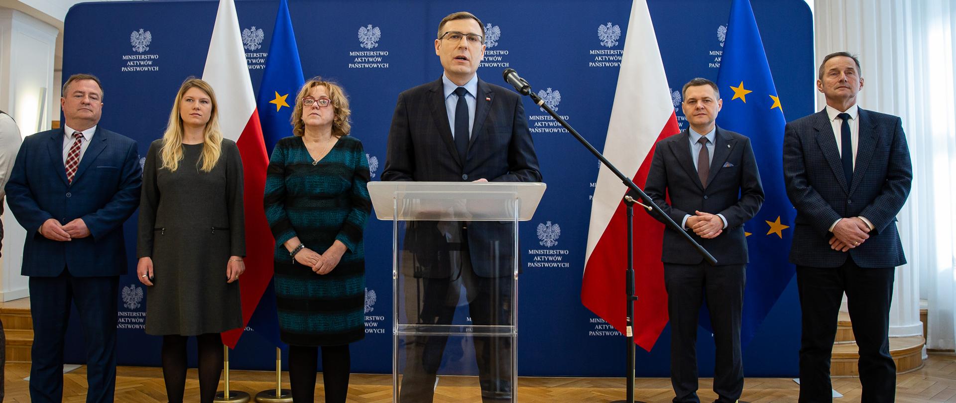 Wiceminister Maciej Małecki stoi przy mównicy. Za nim stoją pozostali uczestnicy briefingu. W tle ścianka z logotypami MAP i flagi. 