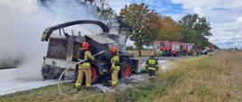 Zdjęcie drogę krajową na której stoi palący się kombajn do koszenia kukurydzy oraz strażaków podczas prowadzenia akcji gaśniczej.