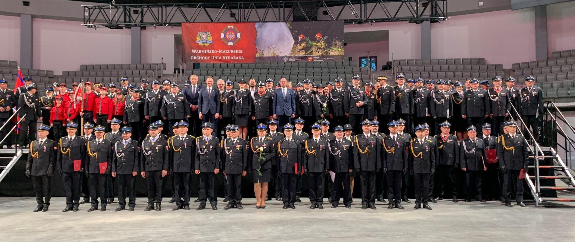 Hala sportowa. Na zdjęciu bardzo duża grupa strażaków PSP i OSP w mundurach galowych. Ustawieni w dwuszeregu na ziemi i na podwyższeniu.