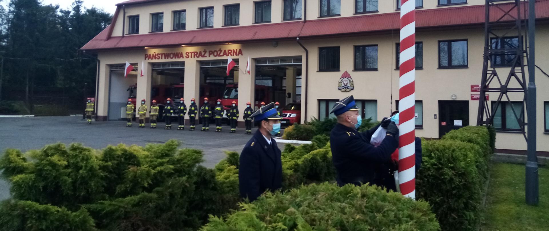 Zdjęcie przedstawia przypięcie flagi państwowej do masztu przez poczet flagowy. W tle budynek Komendy Powiatowej Państwowej Straży Pożarnej w Łańcucie.