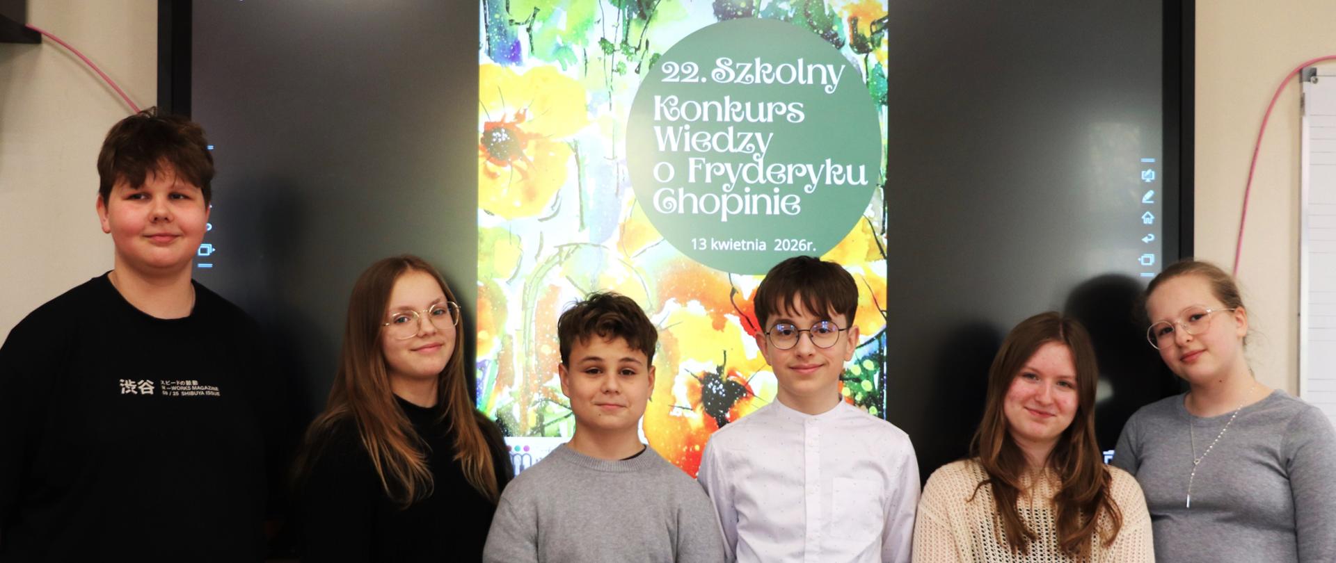 Na zdjęciu widać sześcioro uczniów stojących obok siebie w sali lekcyjnej lub szkolnej. Za nimi znajduje się duży ekran z kolorowym plakatem informującym o „22. Szkolnym Konkursie Wiedzy o Fryderyku Chopinie”, który odbył się 13 kwietnia 2026 roku.
Uczniowie stoją w rzędzie, patrząc w stronę aparatu i lekko się uśmiechając. Są ubrani w codzienne, schludne stroje – swetry, koszule i bluzki. Tło plakatu jest jasne i kwiatowe, co kontrastuje z ciemniejszym ekranem po bokach.