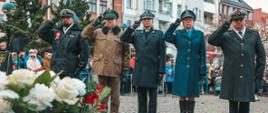 Na zdjęciu widoczni przedstawiciele służb mundurowych stojący w szeregu oddając honor frontem do zdjęcia. Od lewej Marynarka Wojenna, Straż Graniczna, Państwowa Straż Pożarna, Policja, Wojsko Polskie. W tle widoczni inni zgromadzeni. Na pierwszym planie wieniec z białymi kwiatami.