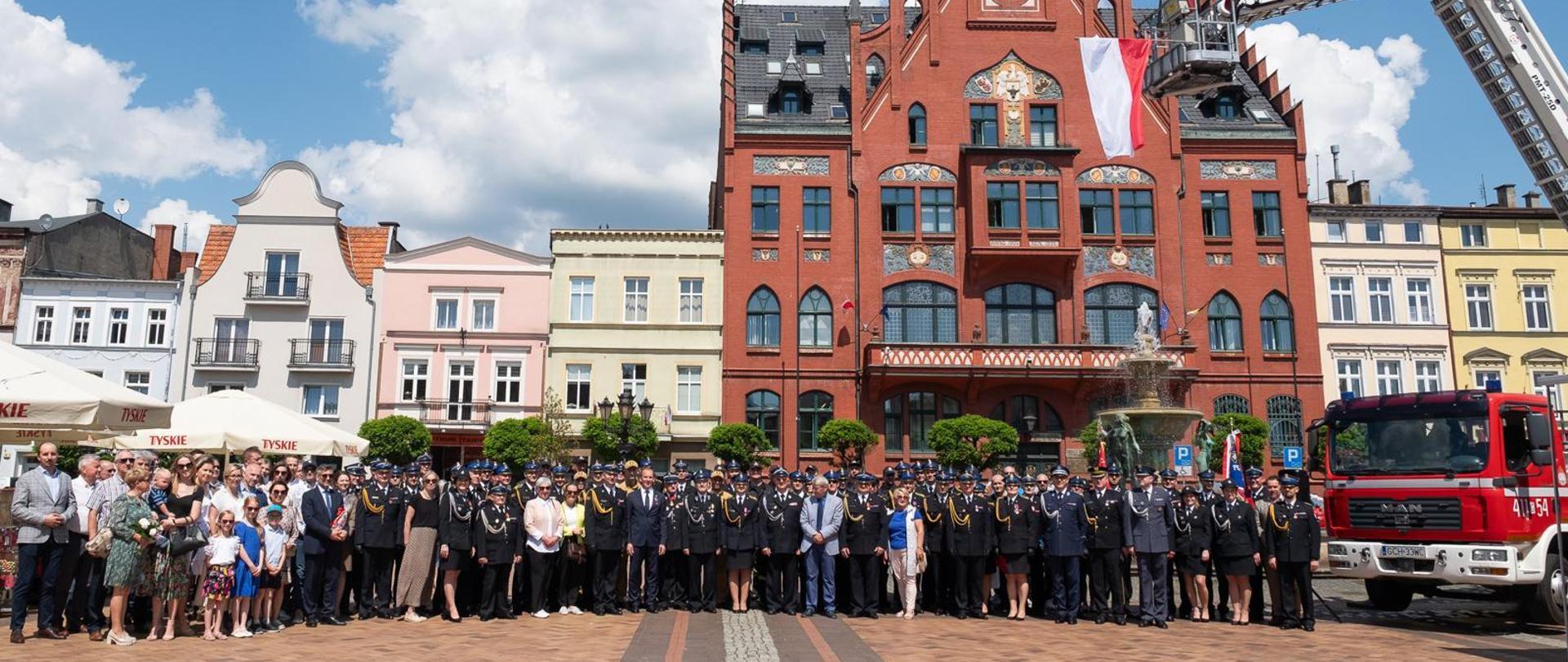 Zdjęcie zbiorowe uczestników uroczystości na płycie Starego Rynku w Chojnicach na tle ratusza. Z prawej strony podnośnik na którym powiewa flaga Polski.