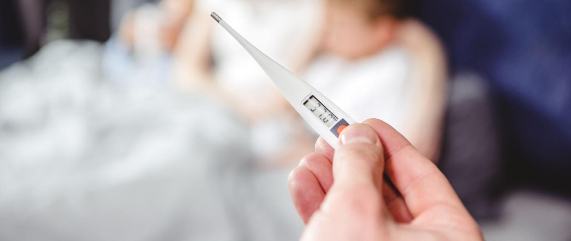 Na zdjęciu jest termometr trzymany w dłoni. W tle jest widoczna kobieta z dzieckiem. Leżą oni w łóżku.