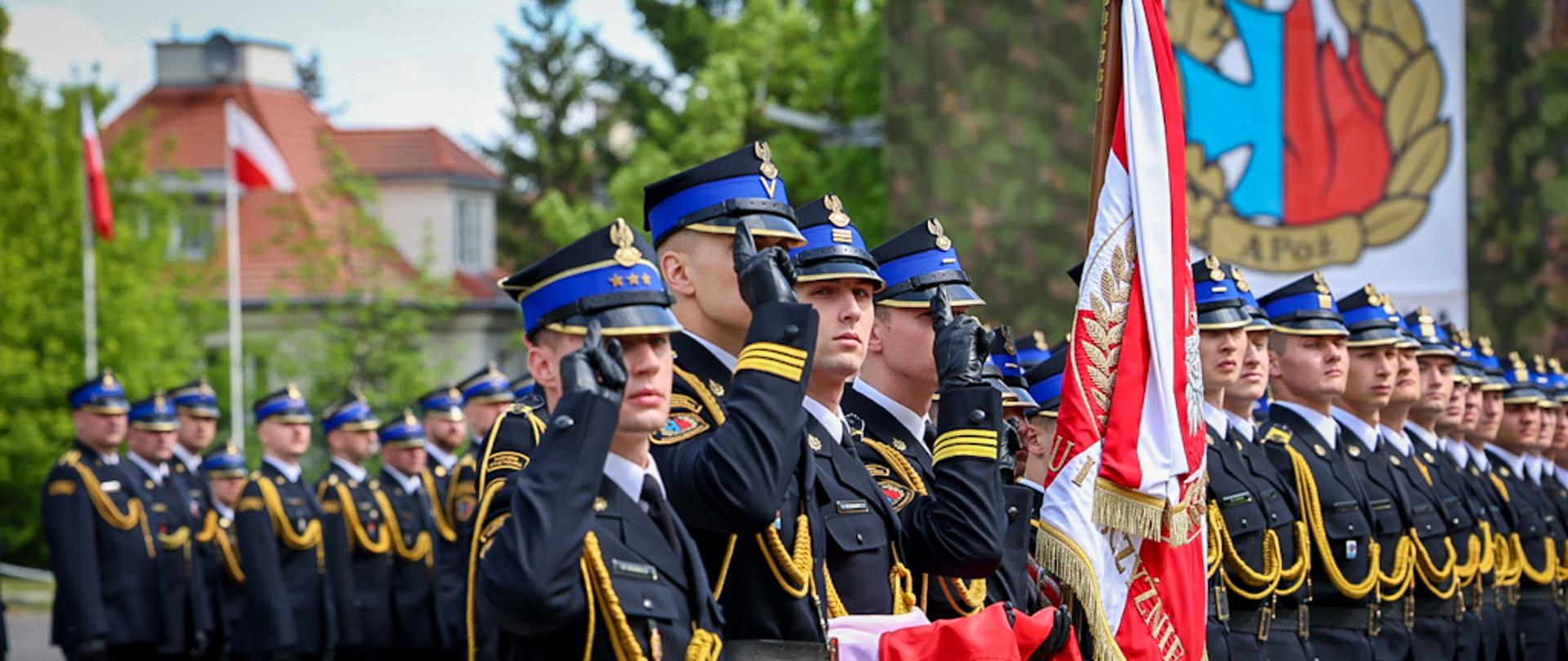 Kompania honorowa Akademii Pożarniczej stoi w szyku podczas uroczystego apelu. Na pierwszym planie poczet sztandarowy, a strażacy w mundurach galowych oddają honory.
