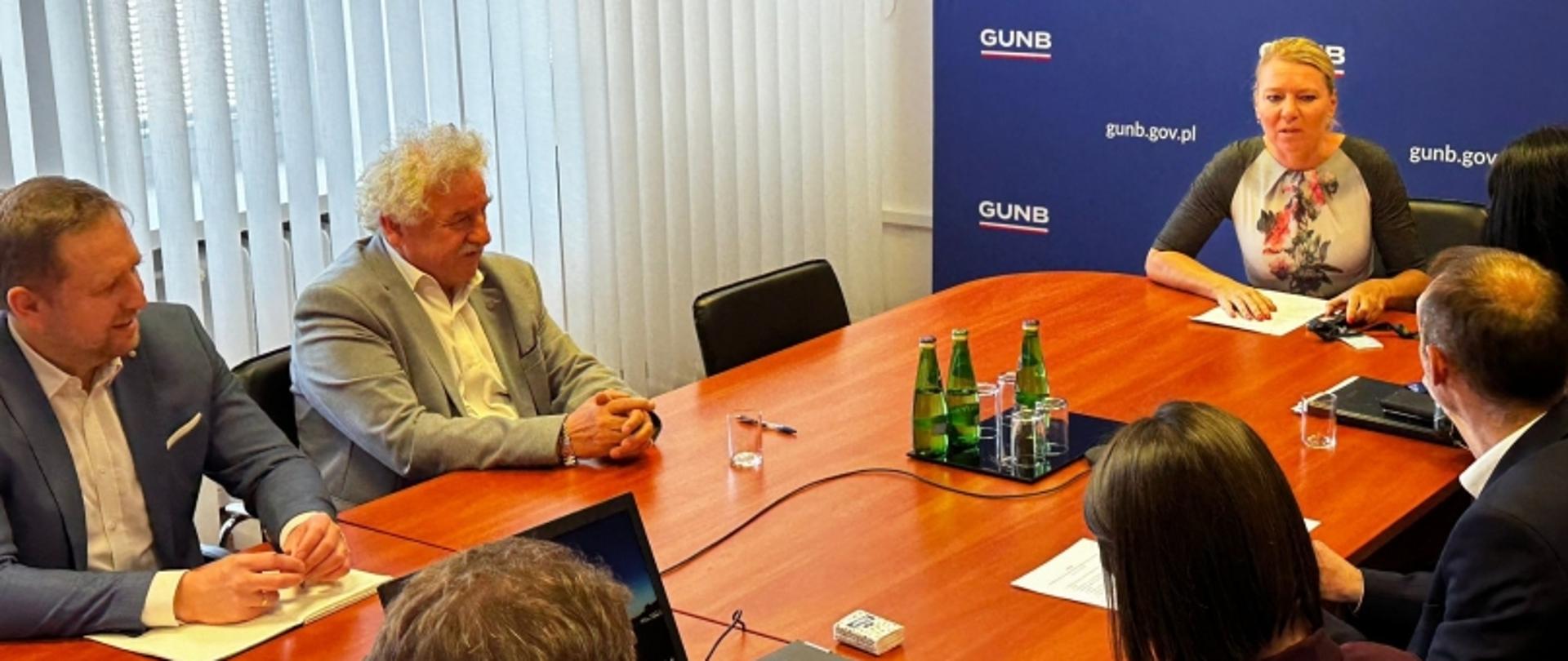 Sześć osób przy stole konferencyjnym, w tle niebieski baner z napisem „GUNB” i adresem „gunb.gov.pl”; na stole zielone butelki, dokumenty i zeszyty.
