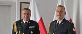 Pomorski komendant wojewódzki Państwowej Straży Pożarnej stoi obok strażaka trzymającego czerwoną teczkę za nimi stoją dwie flagi polski wisi obraz oraz znajdują się drzwi.