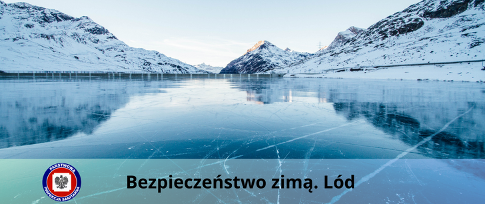 Górzyste pokryte śniegiem wybrzeże nad skutym lodem jeziorem, na dole przezroczysty pasek z czarnym napisem Bezpieczeństwo zimą. Lód a z lewej strony logo Państwowej Inspekcji Sanitarnej.