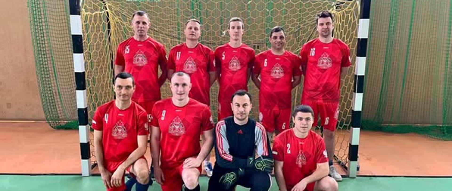 Drużyna strażaków Państwowej Straży Pożarnej w czerwonych strojach sportowych pozuje do wspólnego zdjęcia przed bramką w hali sportowej.