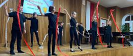 fotografia przedstawia dzieci na scenie, ubrane na czarno, w dłoniach trzymają czerwone oraz białe wstęgi