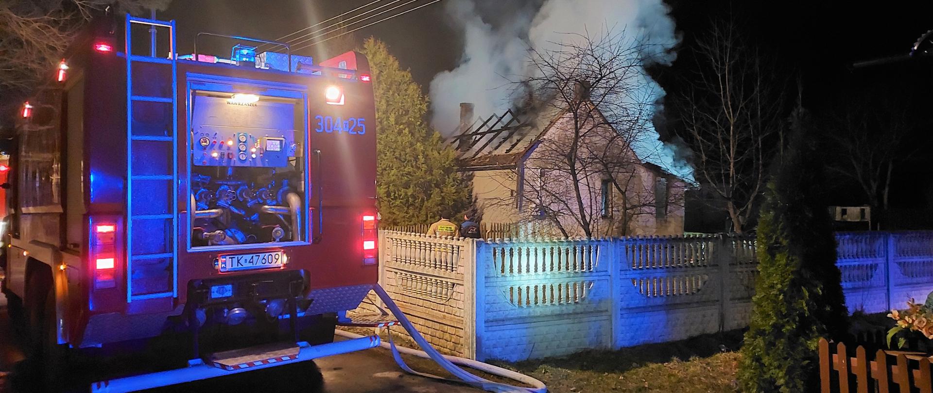 Na zdjęciu przedstawiono: jednorodzinny budynek mieszkalny (po prawej stronie) gdzie wybuchł pożar i pozostawił jedynie zwęgloną konstrukcje dachową. Po lewej stronie stoi samochód pożarniczy.