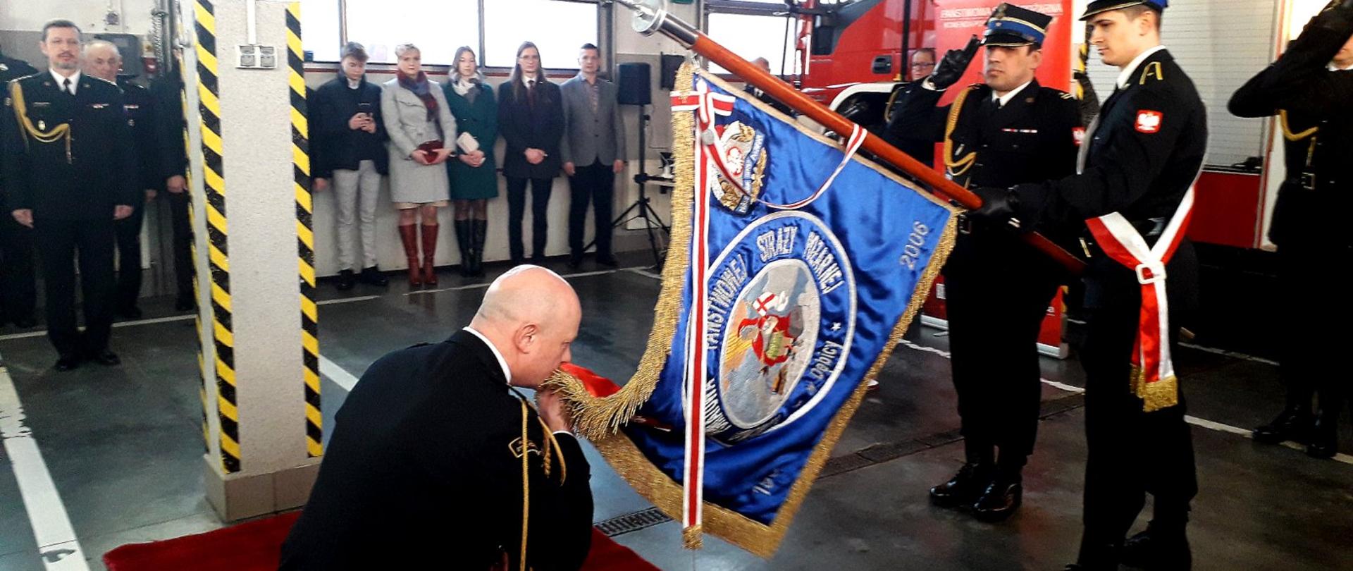 Komendant Powiatowy Państwowej Straży Pożarnej klęcząc na kolanie całuje sztandar Komendy Powiatowej Państwowej Straży Pożarnej w Dębicy. W tle zaproszeni goście i strażacy którzy są ubrani w mundury wyjściowe. Wszyscy znajdują się w pomieszczeniu garażowym straży pożarnej, przy czym w tym momencie znajduje się jeden samochód wewnątrz garażu.