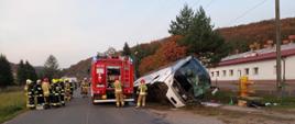 Kolorowa fotografia wykonana w dzień przedstawia widok akcji ratowniczej. W przydrożnym rowie leży na boku, z uszkodzonym przodem, biały autobus. Przy nim na drodze stoi tyłem czerwony, ciężarowy samochód pożarniczy. Przy nim stoi wielu strażaków w ubraniach bojowych z hełmami na głowach. W tle widoczna część budynku a w oddali korony drzew w kolorach jesieni.