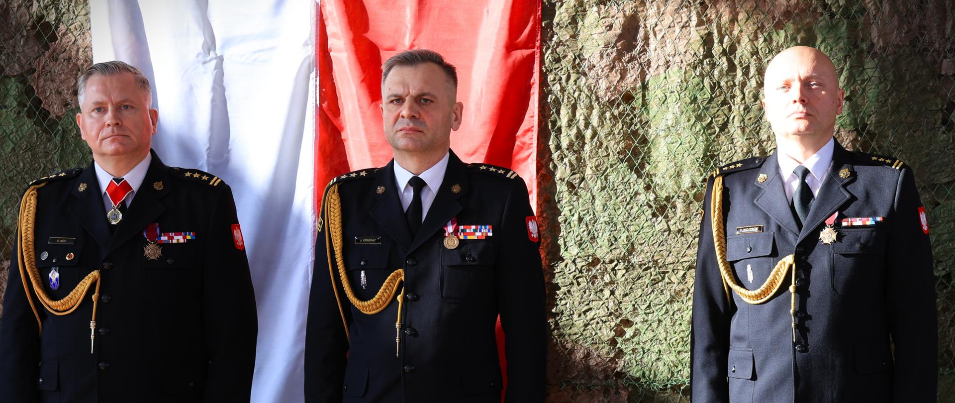 Zdanie i przyjęcie obowiązków Komendanta Miejskiego Państwowej Straży Pożarnej w Białymstoku - na zdjęciu dotychczasowy komendant miejski PSP w Białymstoku, podlaski komendant wojewódzki PSP i nowy komendant miejski PSP w Białymstoku, stoją przy ścianie, w tle barwy narodowe