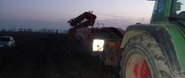 Zdjęcie przedstawia zielony ciągnik rolniczy z podłączoną czerwoną maszyną do zbioru warzyw, która znajduję się na polu.