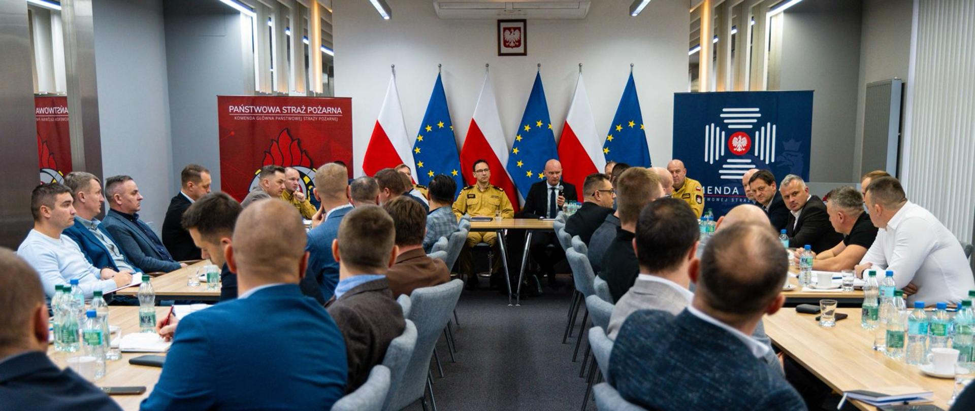 Widok ogólny sali konferencyjnej Komendy Głównej PSP podczas spotkania kierownictwa PSP ze związkami zawodowymi. Uczestnicy siedzą przy długim stole, w tle flagi Polski i Unii Europejskiej.