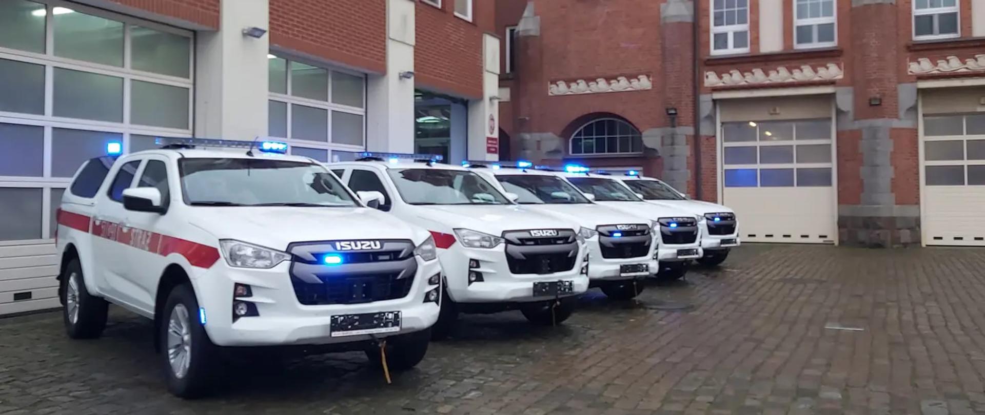 Białe samochody ratownicze Isuzu D-MAX z czerwonymi pasami, bez tablic, na sygnałach świetlnych niebieskich przed garażem Komendy Wojewódzkiej Państwowej Straży Pożarnej. Ustawione są w szeregu, w tle budynek Jednostki ratowniczo gaśniczej nr. 5 w Szczecinie oraz Komenda Wojewódzka PSP w Szczecinie.