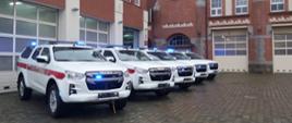 Białe samochody ratownicze Isuzu D-MAX z czerwonymi pasami, bez tablic, na sygnałach świetlnych niebieskich przed garażem Komendy Wojewódzkiej Państwowej Straży Pożarnej. Ustawione są w szeregu, w tle budynek Jednostki ratowniczo gaśniczej nr. 5 w Szczecinie oraz Komenda Wojewódzka PSP w Szczecinie.