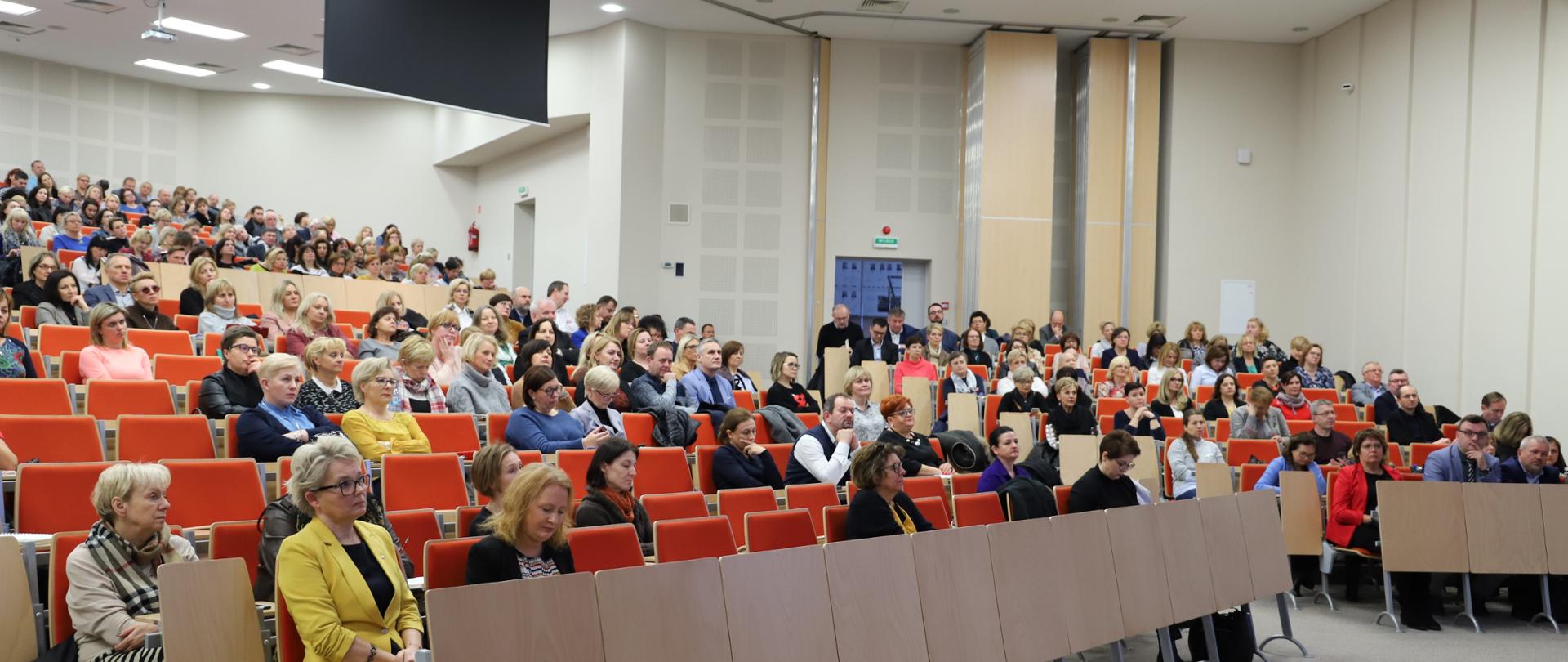 Trwają spotkania informacyjne kuratorów oświaty z dyrektorami szkół w sprawie profilaktyki zdrowotnej. Na dużej sali siedzą dyrektorzy szkół. 