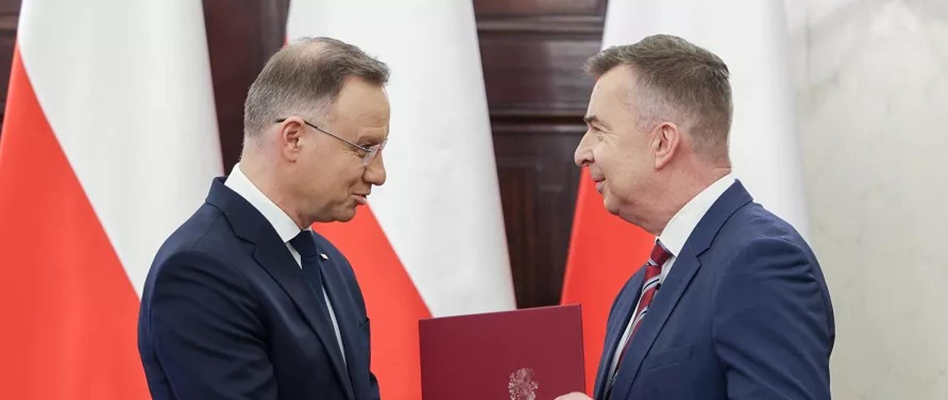 Minister Wieczorek stoi naprzeciwko prezydenta Dudy, podają sobie ręce, za nimi polskie flagi.